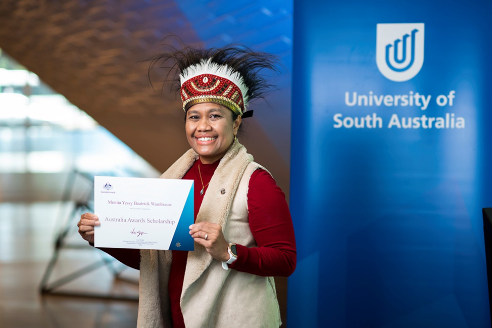 Monita wearing a traditional cassowary feather headdress from her hometown of Biak, Papua with her Australia Awards certificate in UniSA&rsquo;s Pridham Hall.