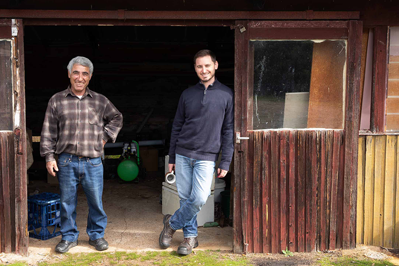 Two olive farmers smile, facing the camera. They are framed in the wide doorway of a rundown looking iron and wood shed. One man holds a china mug