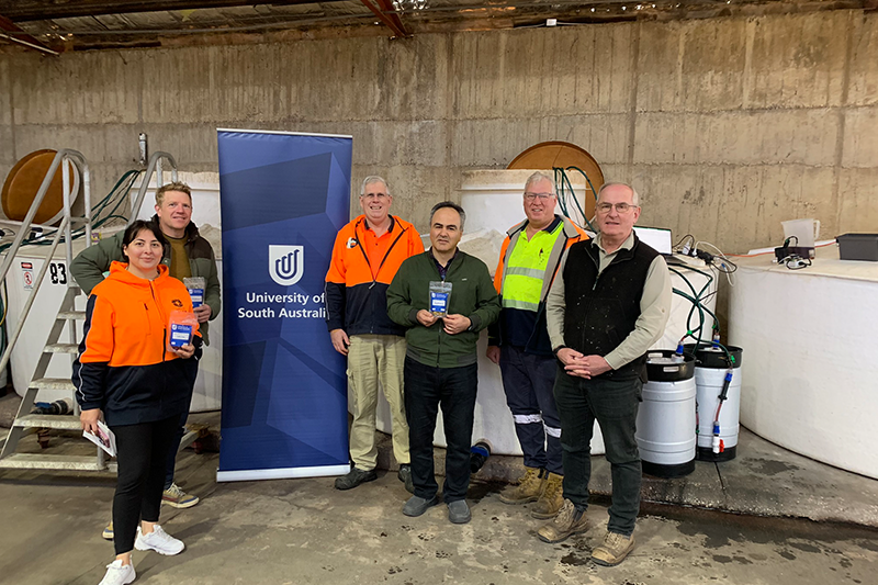 UniSA researchers and collaborators stand in an industrial warehouse in front of a blue UniSA pull-up banner. Some hold little plastic sachets of olives