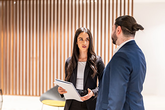 A woman dressed in a business suit speaks to another colleague, also dressed in a suit. They stand near a backdrop of wooden panelling. 