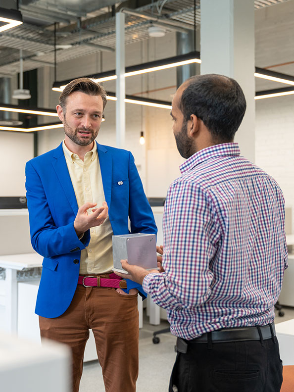 A man in a blue suit jacket faces another man in a light blue shirt. The man in the light blue shirt holds a sillver cube about the size of a child's soccer ball. 