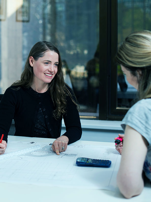 A women in a dark top sits a desk with a large paper blue print  on the table in front of her. She is talking to another person who is almost out of frame. 
