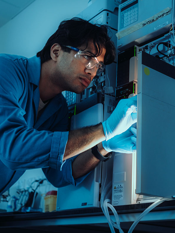 A man with a blue lab coat and light blue plastic gloves inspects a machine. The machine is casting a yellow glow on his face. 