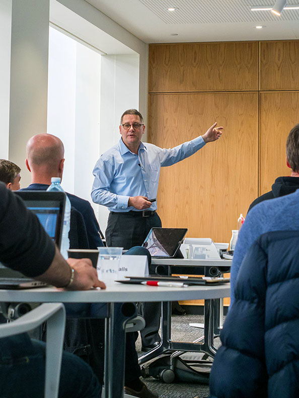 A man in a light blue shirt presents to a room full of people. The crowd's backs are facing away from the camera. 